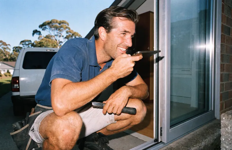 Smiling Australian tradesperson speaking on a phone while fitting a sliding window